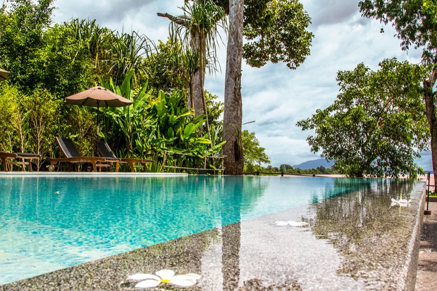An infinity pool edged with sun loungers, framed by palms, with the Mekong and low hills beyond