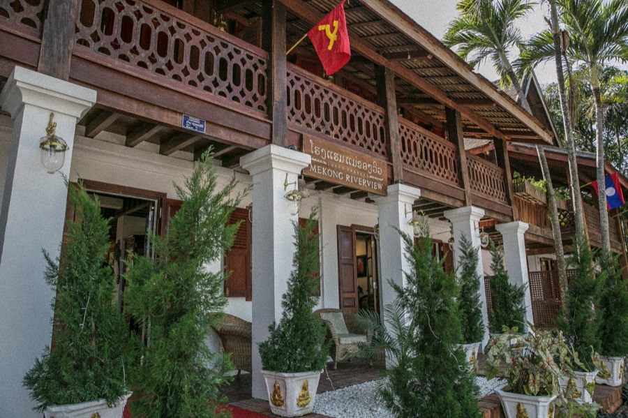 The front of a two-storey wooden and white-painted building with a carved upper balcony and palms to the side