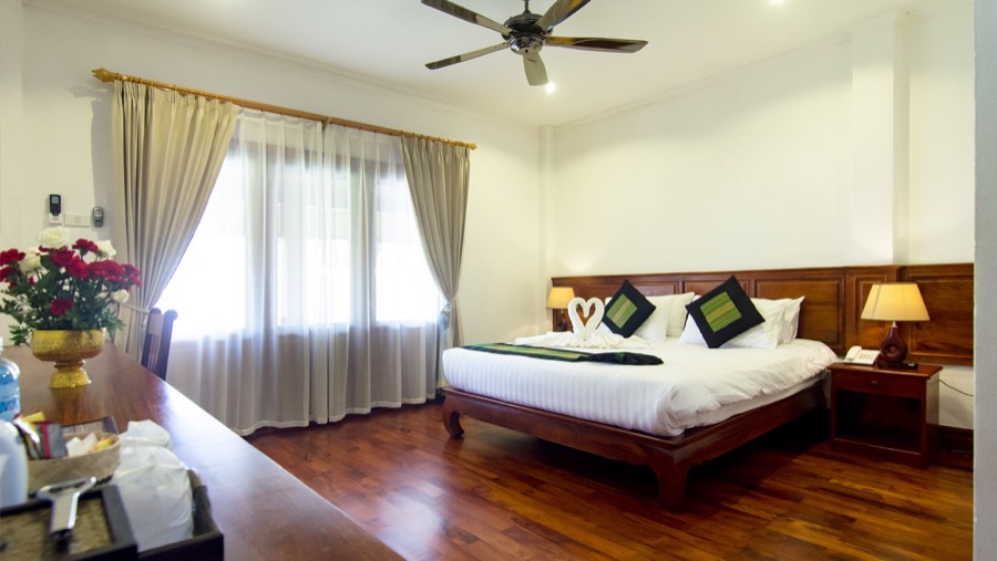A room with a wooden double bed, ceiling fan, curtained window and wooden floor
