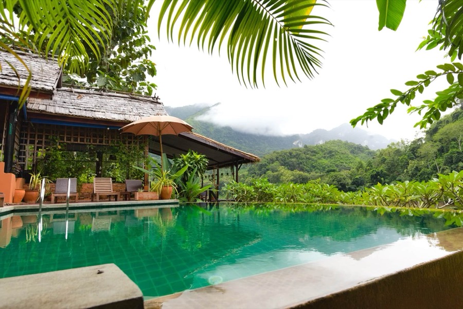 An infinity pool beside a wooden bungalow, with forested limestone hills behind