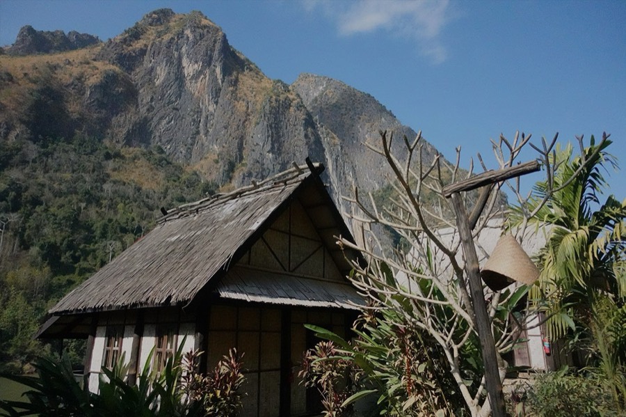 A thatched-roof wooden bungalow among garden plants, with a steep limestone peak behind