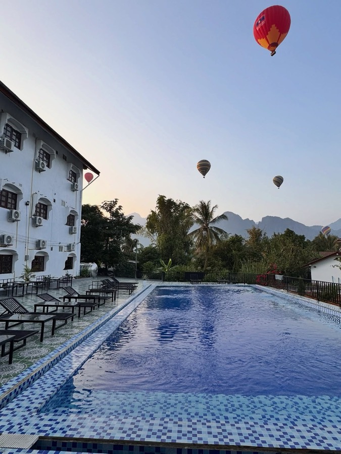 An outdoor pool and sun loungers beside a white guest block, with karst peaks and hot-air balloons behind