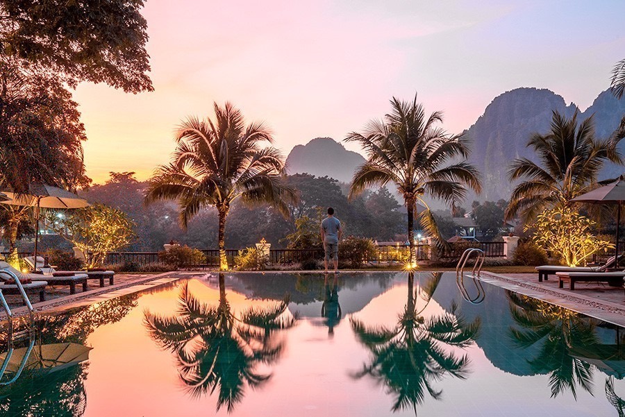 A long pool reflecting palms at dusk, with limestone karst peaks and a pink sky behind